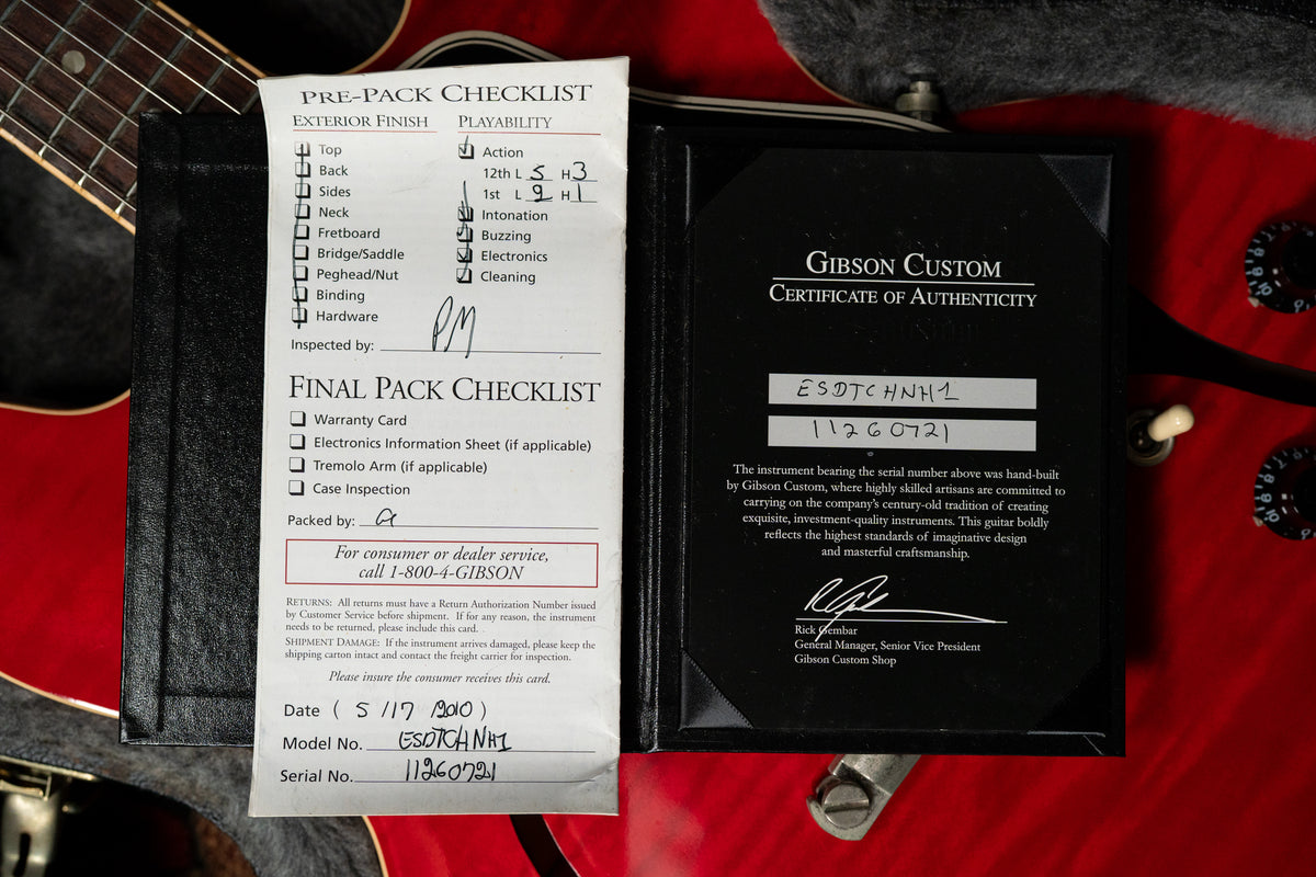 Close-up of a red guitar body with a white "Pre-Pack Checklist" and a black "Certificate of Authenticity" on top.