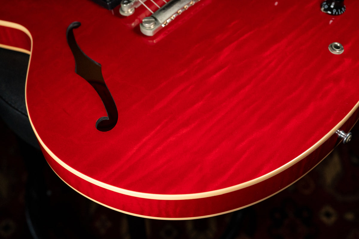 The upper bout of a cherry red flamed maple semi-hollow guitar, showing an f-hole, bridge, and a volume knob.