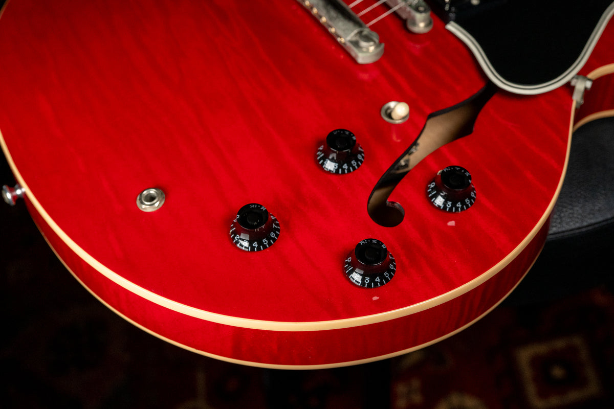 Close-up of the lower bout of a cherry red flamed maple guitar, showing four black numbered control knobs and a cream bound f-hole.