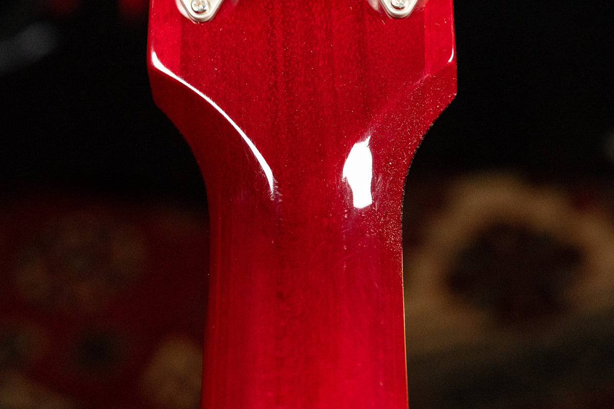 Close-up of the cherry red guitar neck's heel, showing light scratches and dust on the glossy finish, with two tuner posts visible.