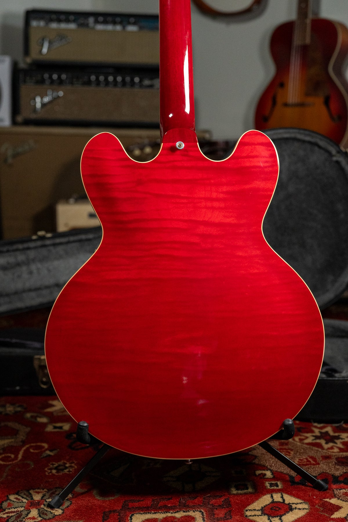 The back of a cherry red flamed maple guitar, showing the neck joint and body binding, on a stand over a patterned rug.