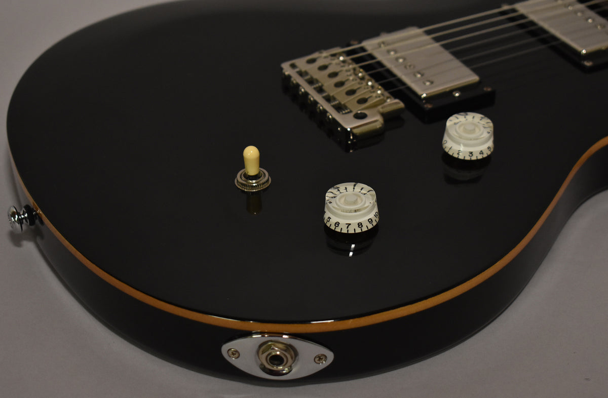 Close-up of a glossy black guitar body with cream binding, showing the output jack, two clear numbered knobs, and a cream toggle switch.