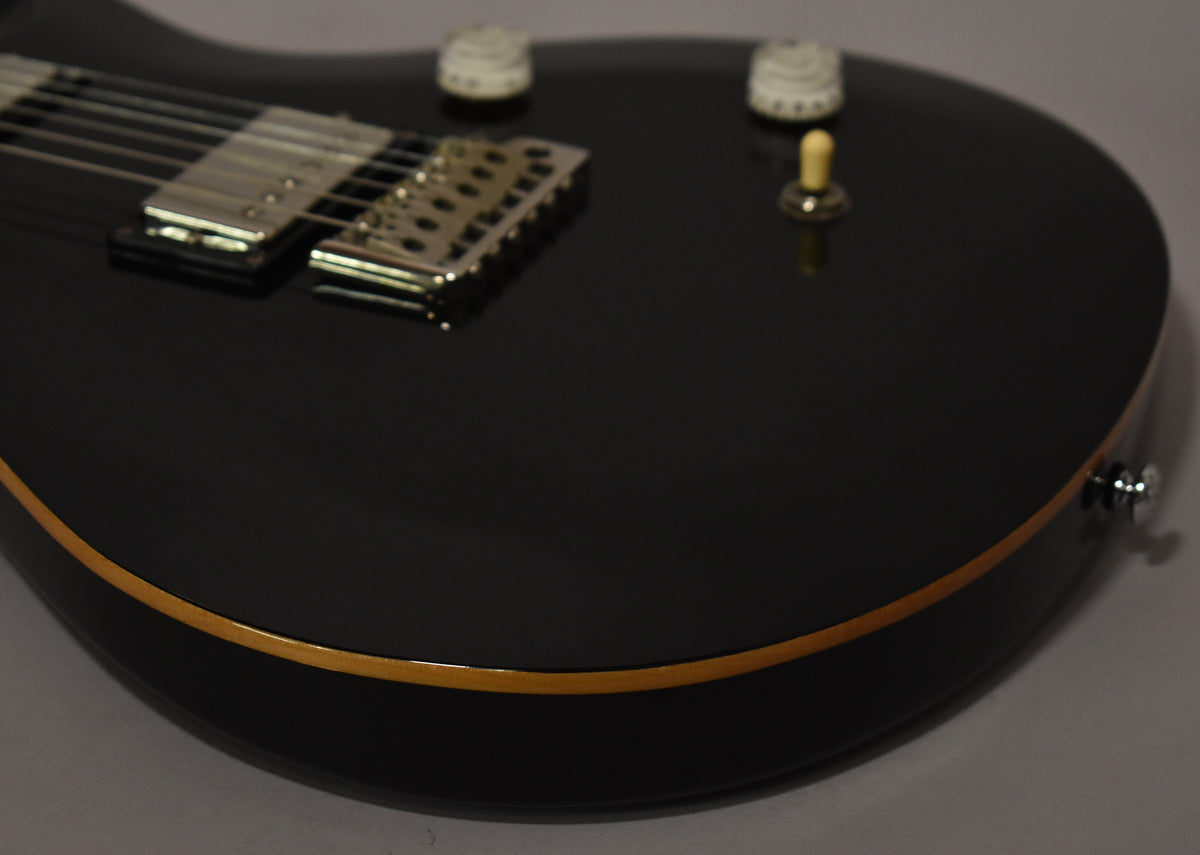Close-up of the black guitar body with natural wood binding, showing chrome bridge, neck pickup, two cream knobs, and a toggle switch.