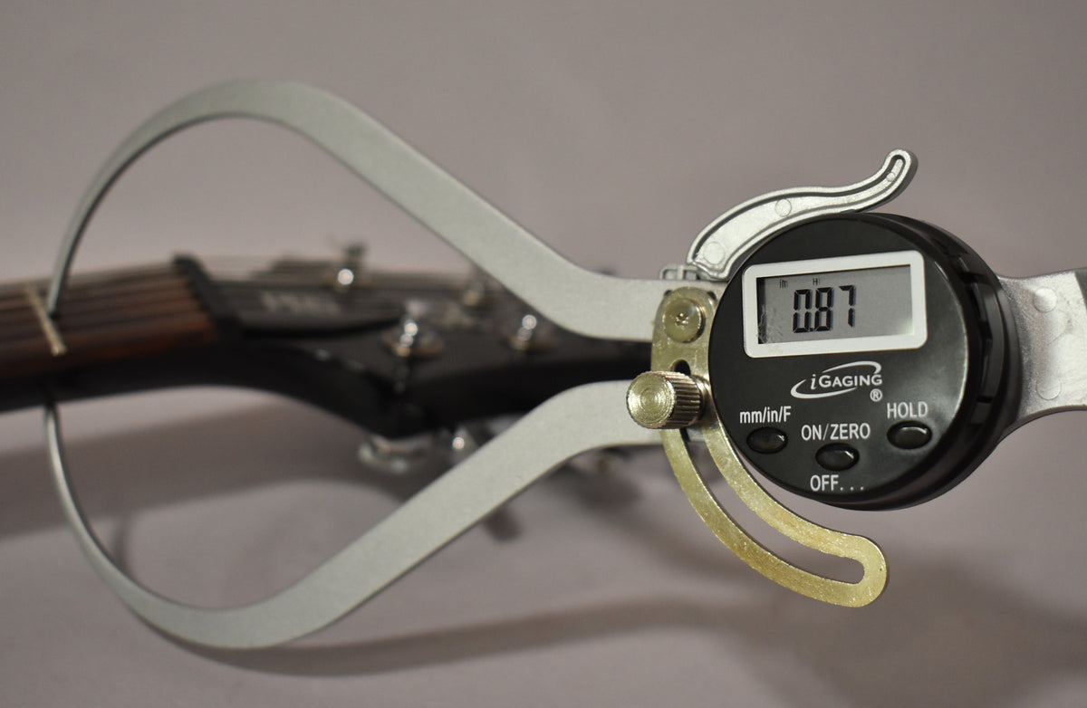 A digital caliper measures the guitar's nut width at 0.87, with the black headstock and dark fretboard visible behind it.
