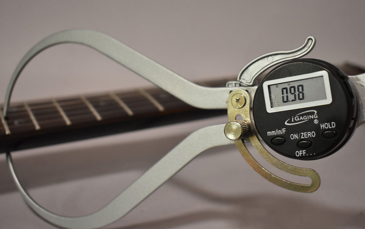 A digital caliper measures the guitar's dark fretboard and frets, displaying "0.98" on its screen.