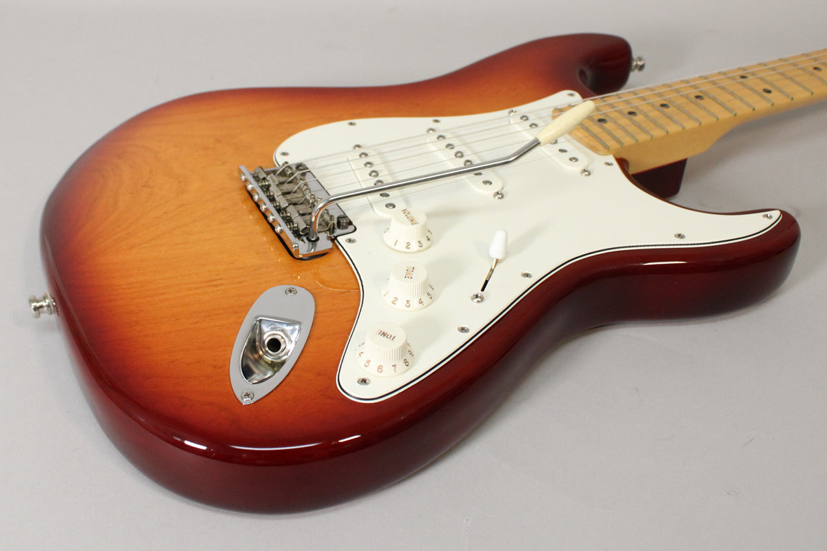 Angled view of a Sienna Burst Stratocaster body, showing the white pickguard, three cream control knobs, and chrome tremolo bridge.