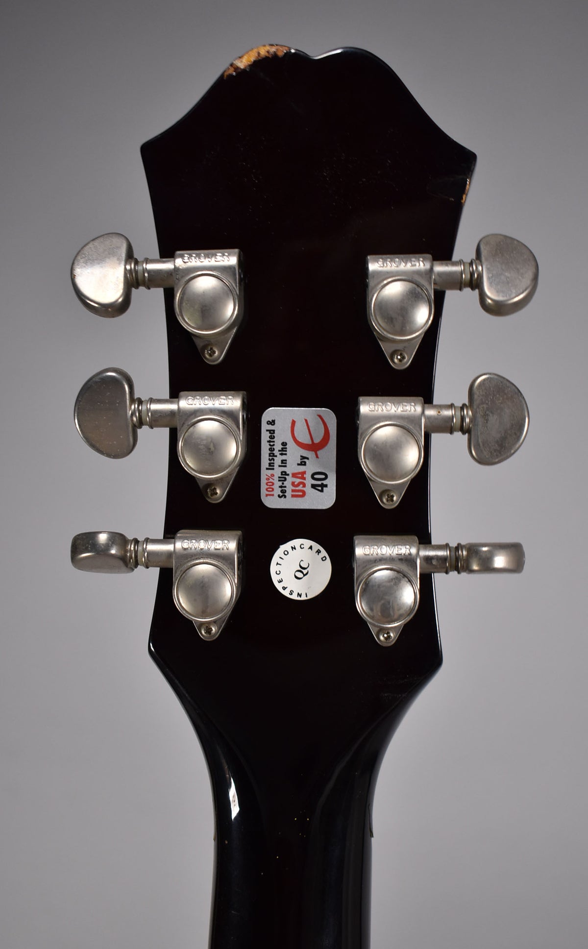 Rear view of the guitar's dark brown headstock with six chrome tuners, two inspection stickers, and paint chipping at the top edge.