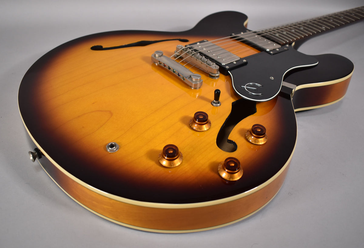 The guitar's lower body, with a sunburst finish, f-hole, four amber control knobs, and a black pickguard, is angled from the left.