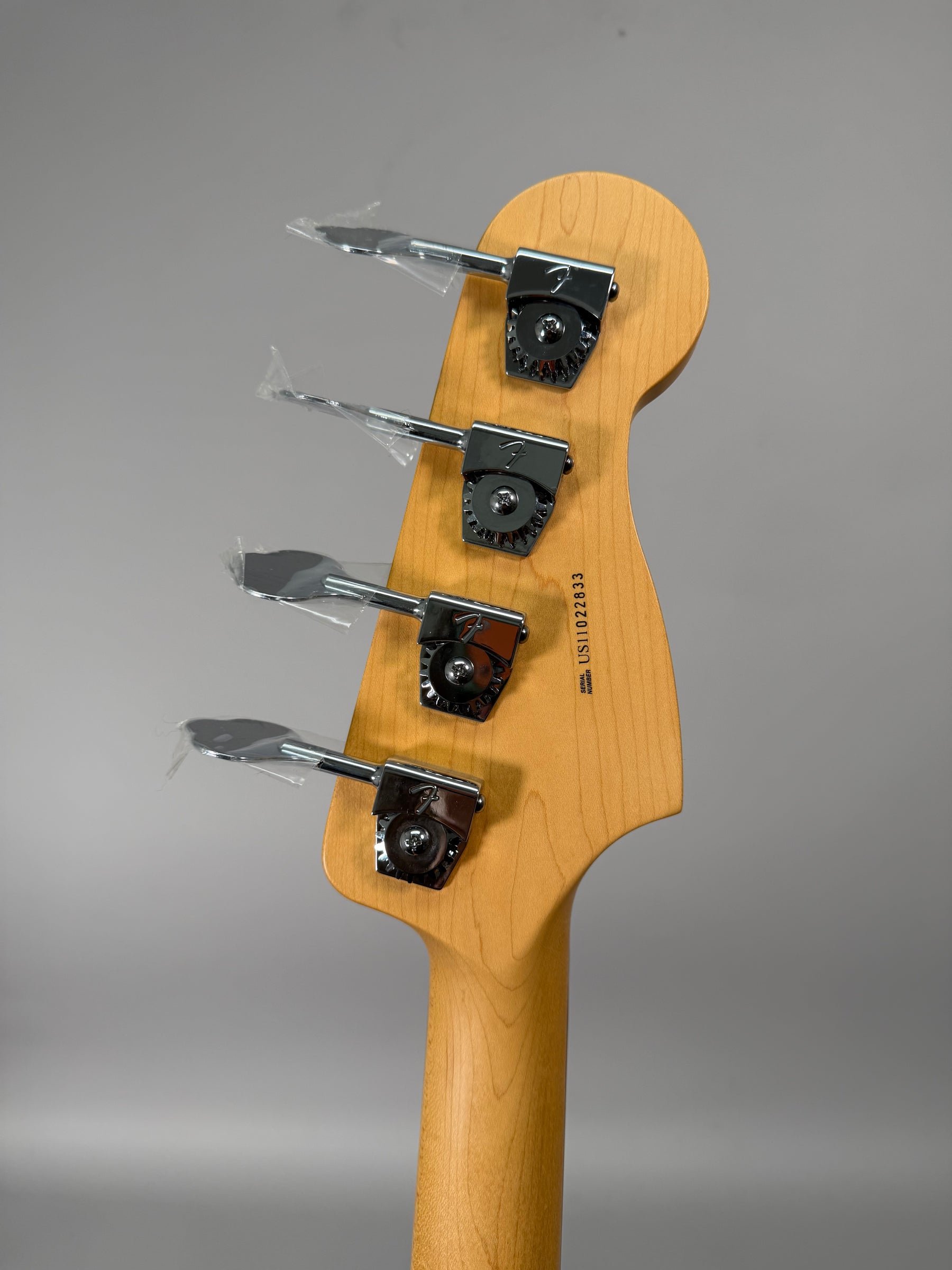 The back of a light maple headstock and neck, with four chrome tuners, each still wrapped in clear plastic.