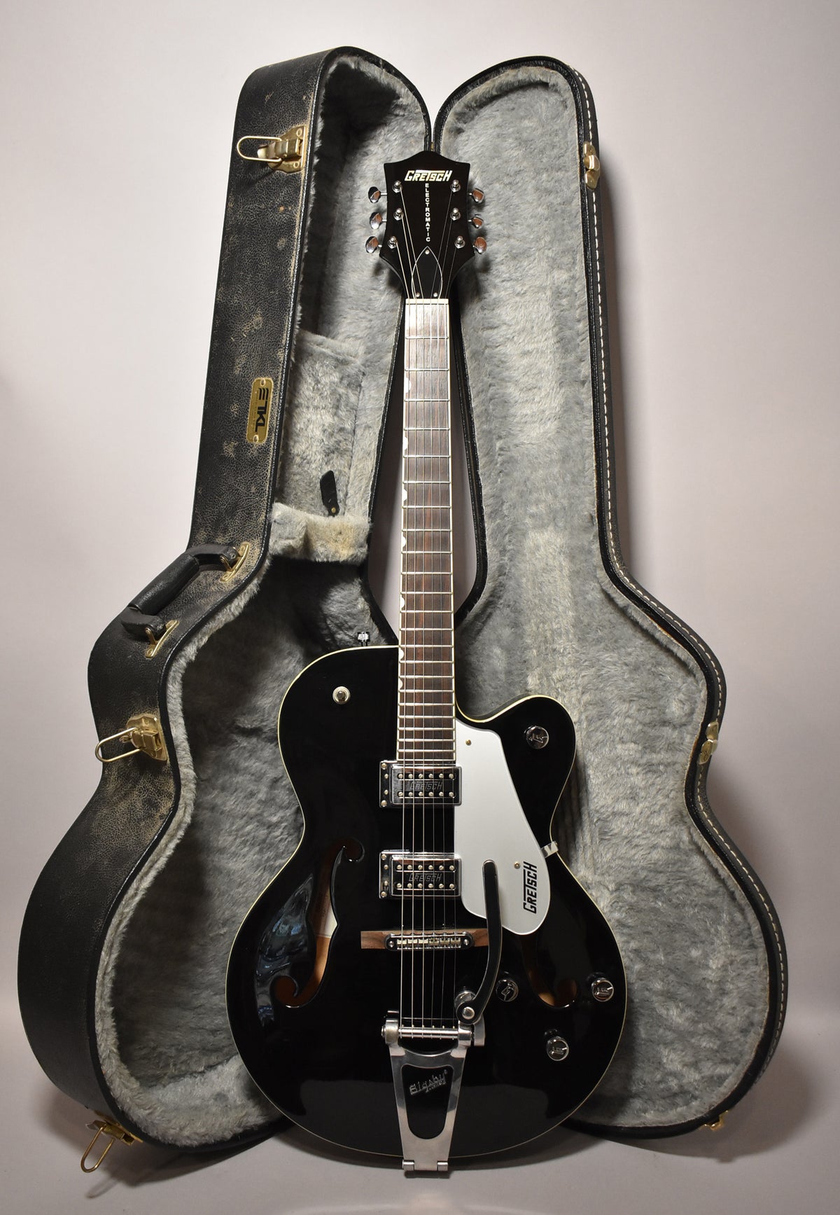 A black hollowbody electric guitar with a white pickguard and Bigsby tremolo stands upright in its open, grey-lined hard case.