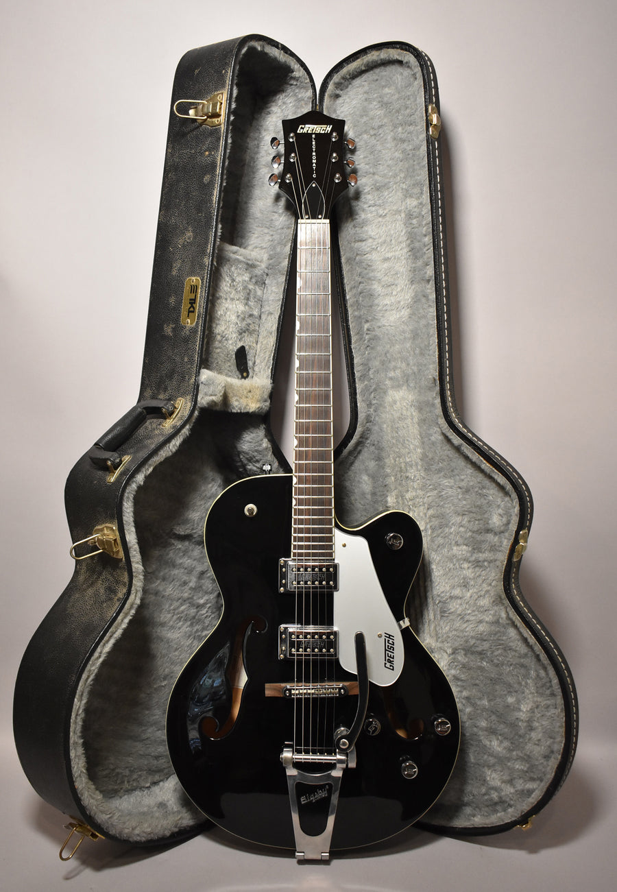 A black hollowbody electric guitar with a white pickguard and Bigsby tremolo stands upright in its open, grey-lined hard case.