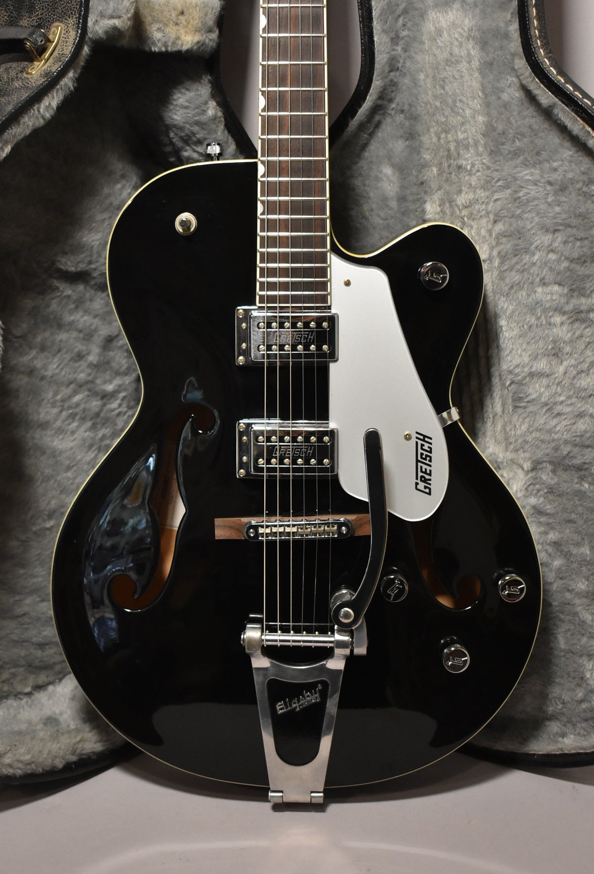 Black hollowbody electric guitar with a white pickguard, two humbuckers, a Bigsby tremolo, and f-holes, shown in a plush case.