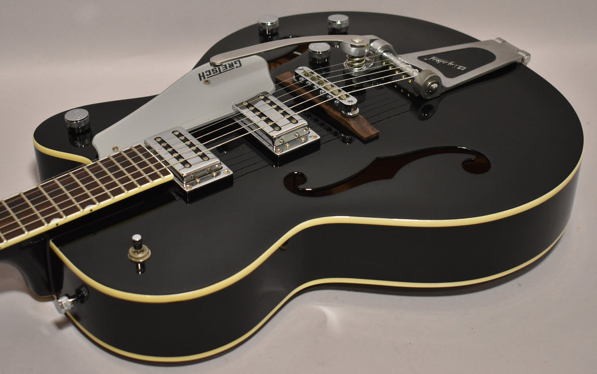 Black hollowbody electric guitar, angled from the lower bout, showing a Bigsby vibrato, two humbuckers, and a white pickguard.