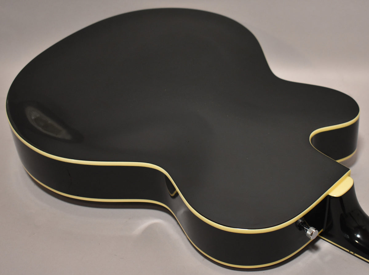 The black hollowbody electric guitar is viewed from the back, showing its matte finish, cream binding, and output jack.
