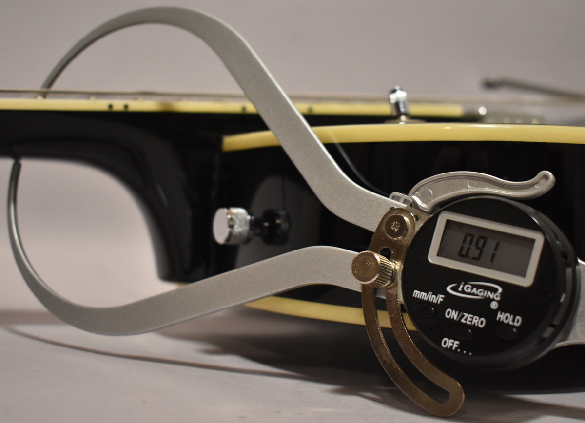 A digital caliper measures the black guitar's neck thickness at 0.91 inches, showing the cream binding and a chrome strap button.