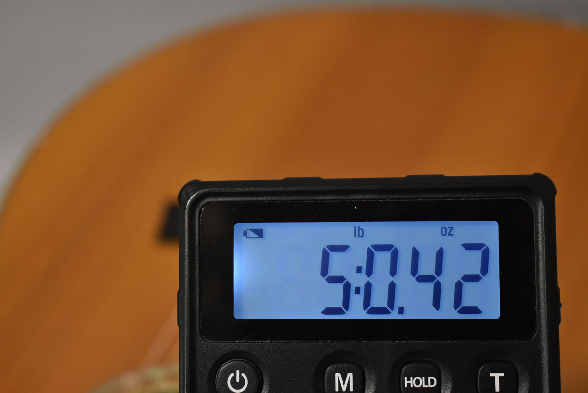 A close-up of a digital scale displaying "5.042 lb" in blue numbers, with a blurred natural wood guitar body in the background.
