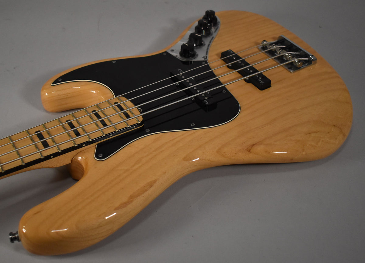 Angled view of a natural finish bass with a black pickguard, two pickups, and a maple fretboard with block inlays.