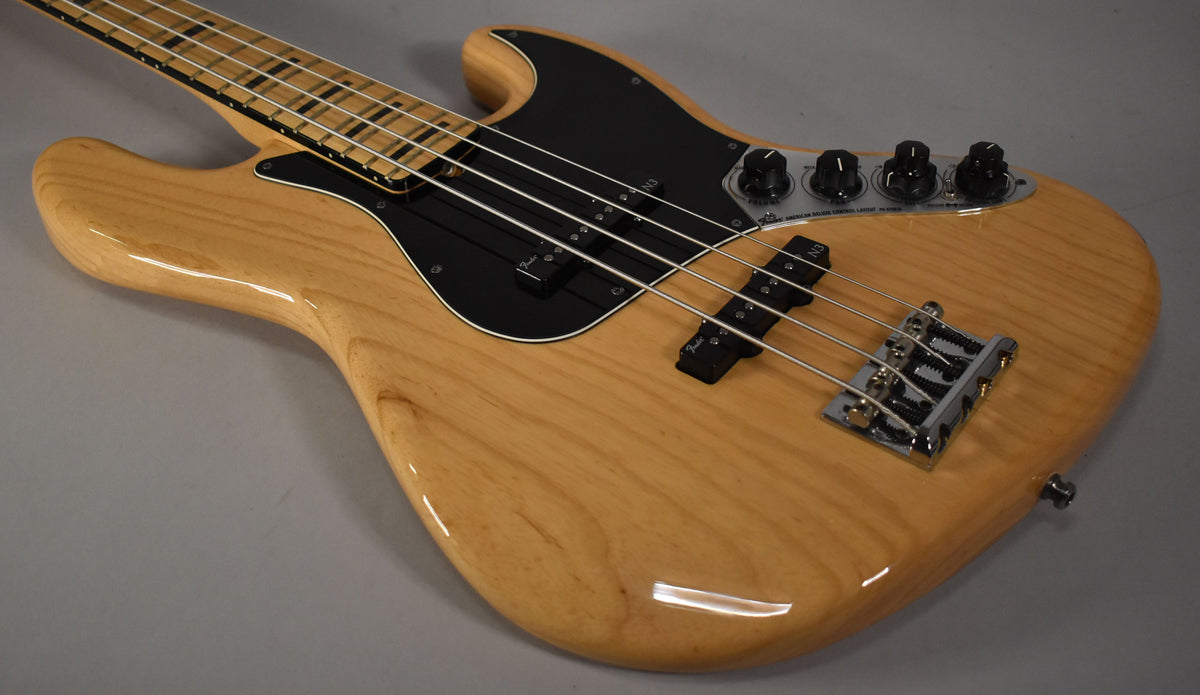 The upper body of a natural finish bass, showing the black pickguard, two pickups, chrome bridge, and four black control knobs.