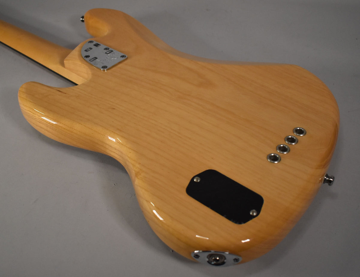 The bass's natural finish body is seen from the back, angled left, showing the neck plate, battery cover, and string ferrules.