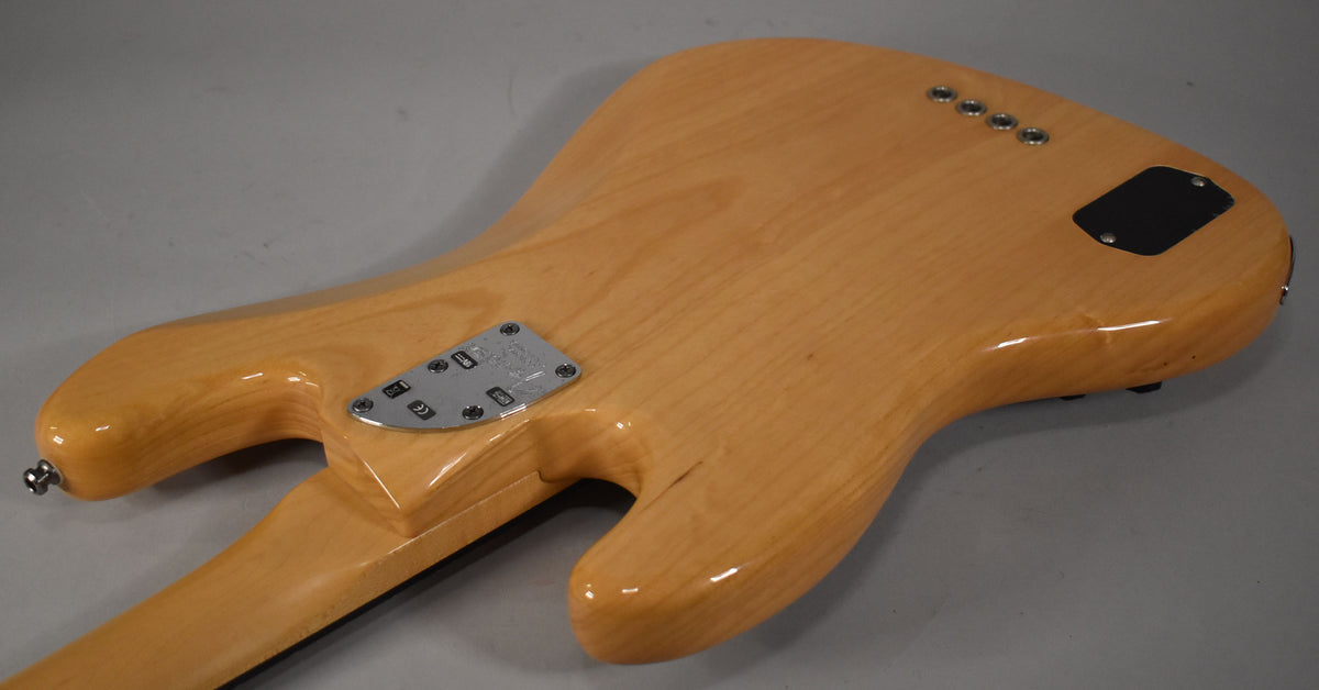 The back of a natural finish bass guitar, angled left, showing the neck plate, black control cavity cover, and four string ferrules.