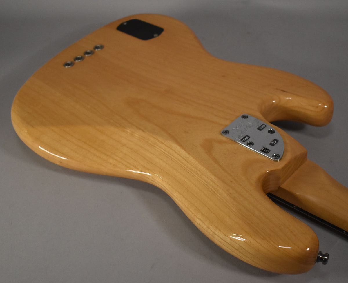 The bass's natural finish body is viewed from the back, showing the neck plate, string ferrules, and a black control cavity cover.