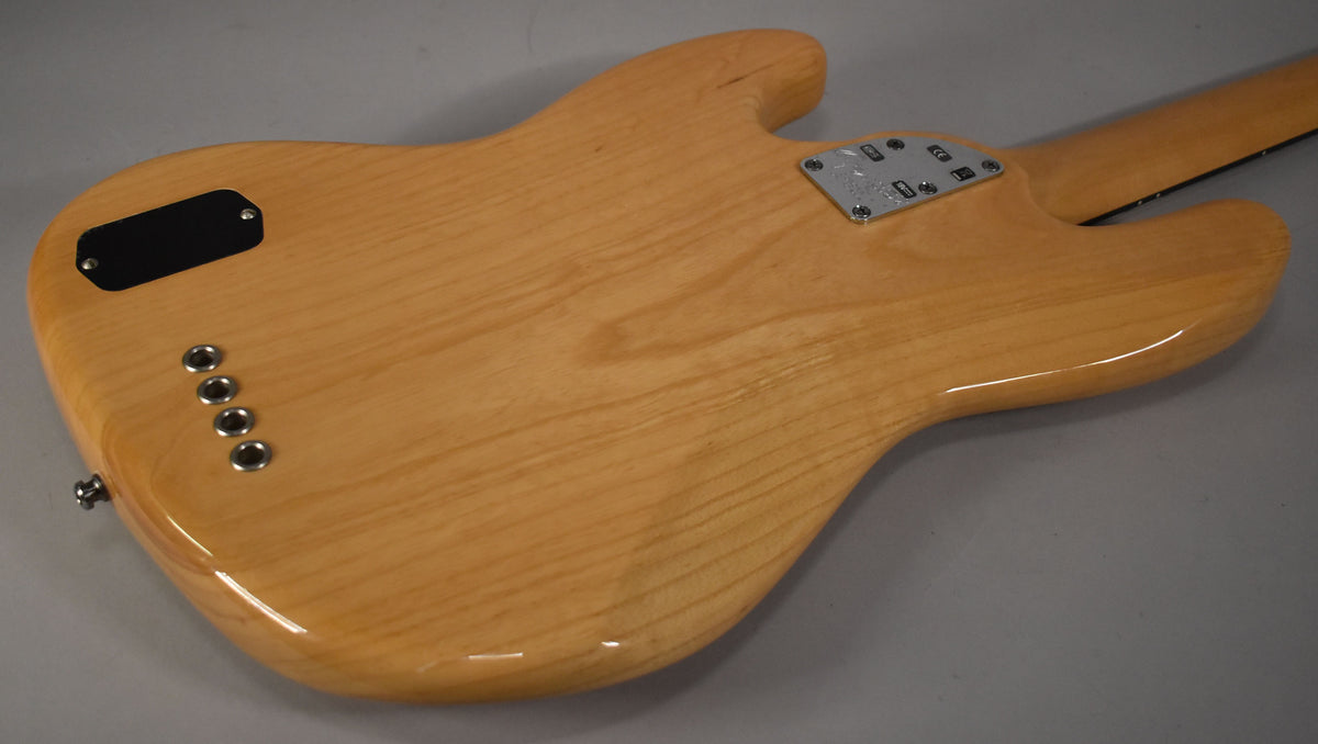 The bass's natural finish body is viewed from the back, showing the neck plate, battery compartment, and four string-through-body ferrules.