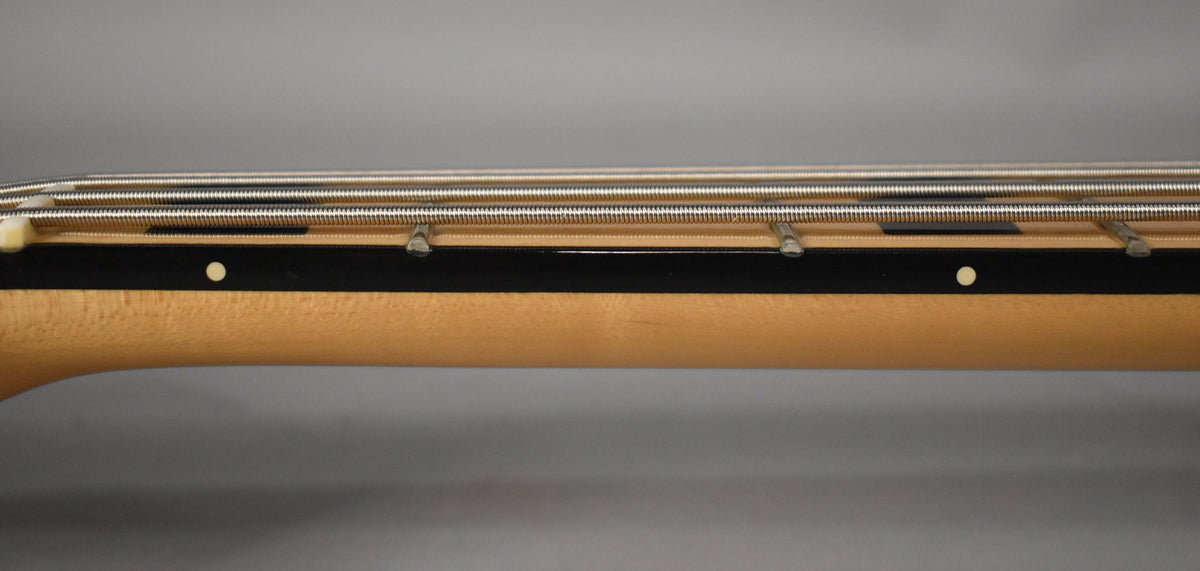 Close-up of the bass neck, showing the light maple wood, dark fretboard with white dot inlays, and four silver strings.