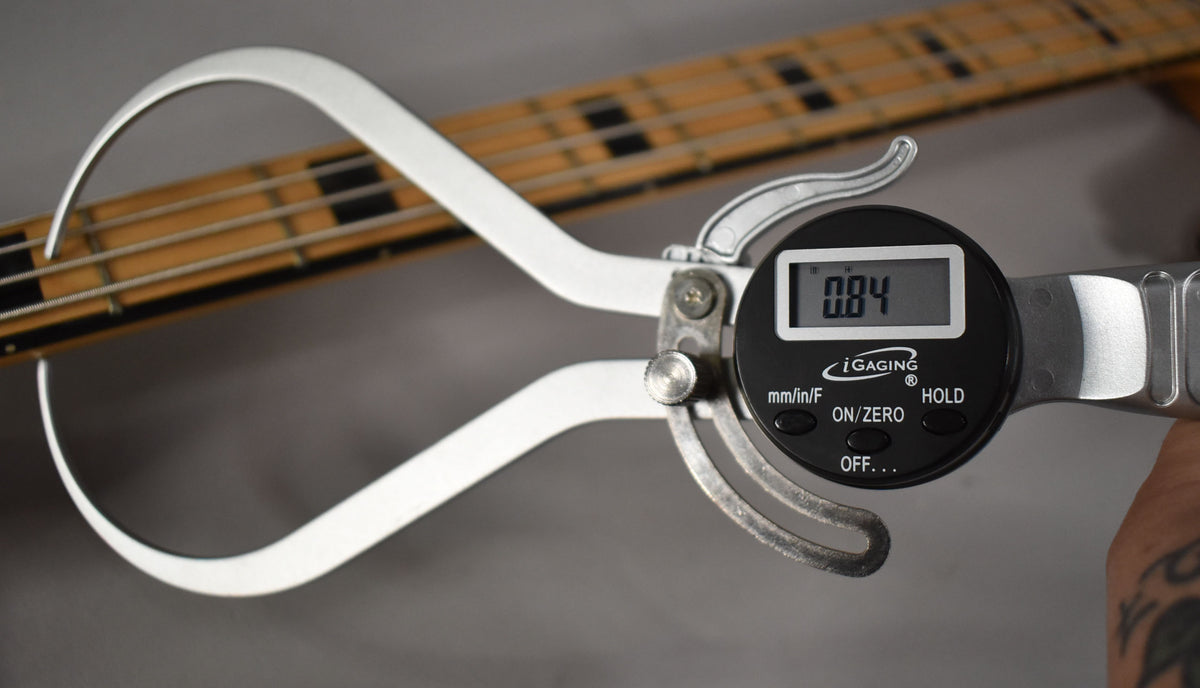 A close-up of a digital caliper measuring the bass's fretboard, showing a reading of 0.84, with a maple neck and block inlays visible.