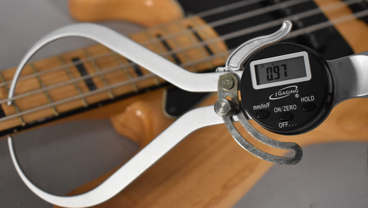 A close-up of the bass's natural finish maple fretboard, with a digital caliper measuring the string height at 0.97.