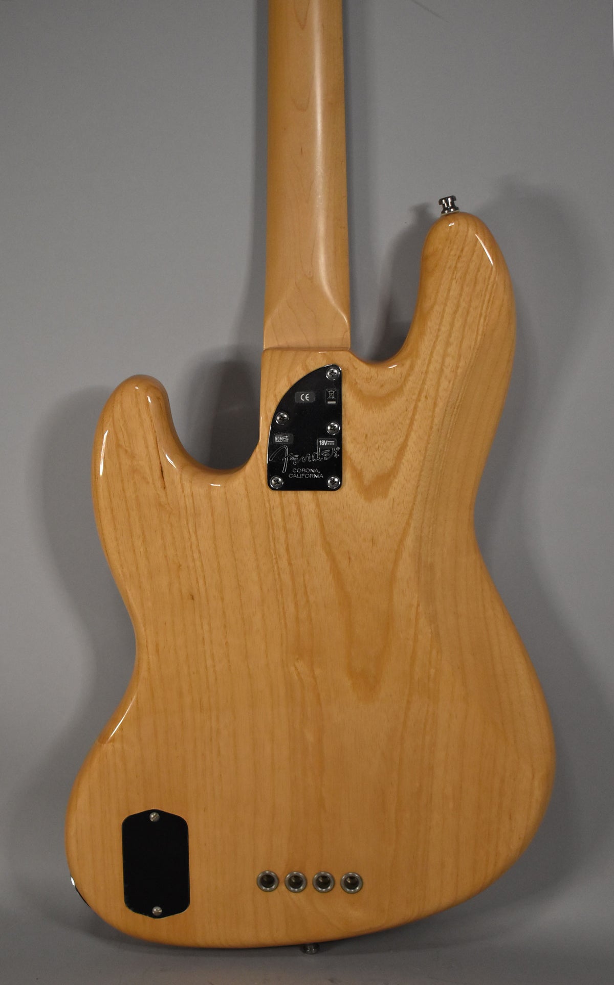 The back of a natural finish bass, showing the neck joint, a black battery compartment, and four chrome control knobs.