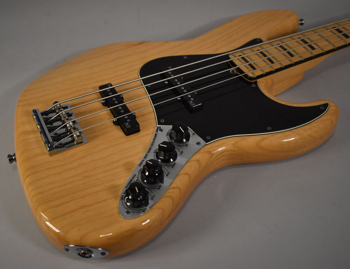 Natural finish Jazz Bass, angled from the lower bout, showing the chrome bridge, black pickguard, and control plate with four black knobs.