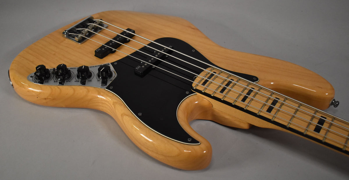 Natural finish bass, angled from the lower bout, showing the black pickguard, control plate with four black knobs, and maple fretboard.