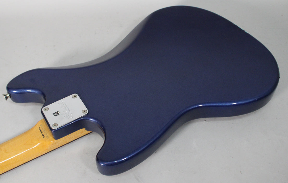 The back of a metallic blue guitar body, angled from the lower left, showing the neck plate and a portion of the light-colored neck.