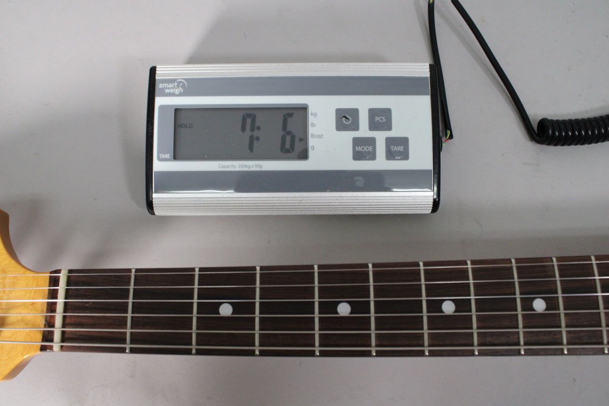 The guitar's dark fretboard and light maple neck are visible below a digital scale displaying "7.6" lbs.