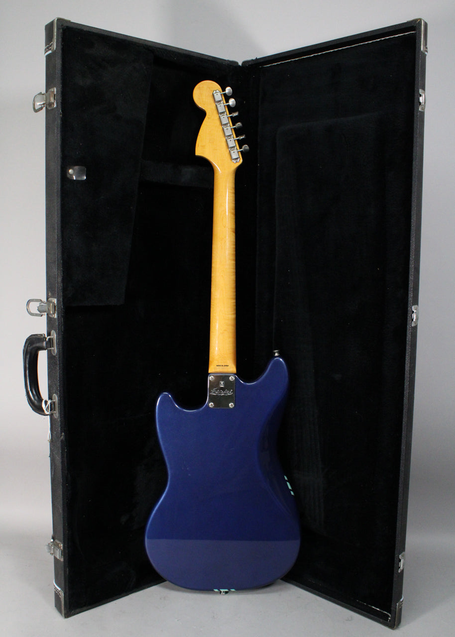 The back of a glossy blue electric guitar with a light maple neck and headstock, standing in an open black hard case.
