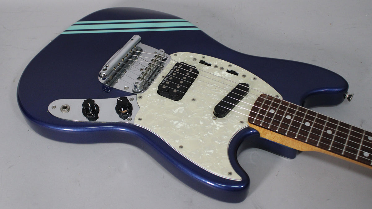 Dark blue electric guitar, angled from above, showing the body with two light blue racing stripes, white pearloid pickguard, and black humbucker and single-coil pickups.