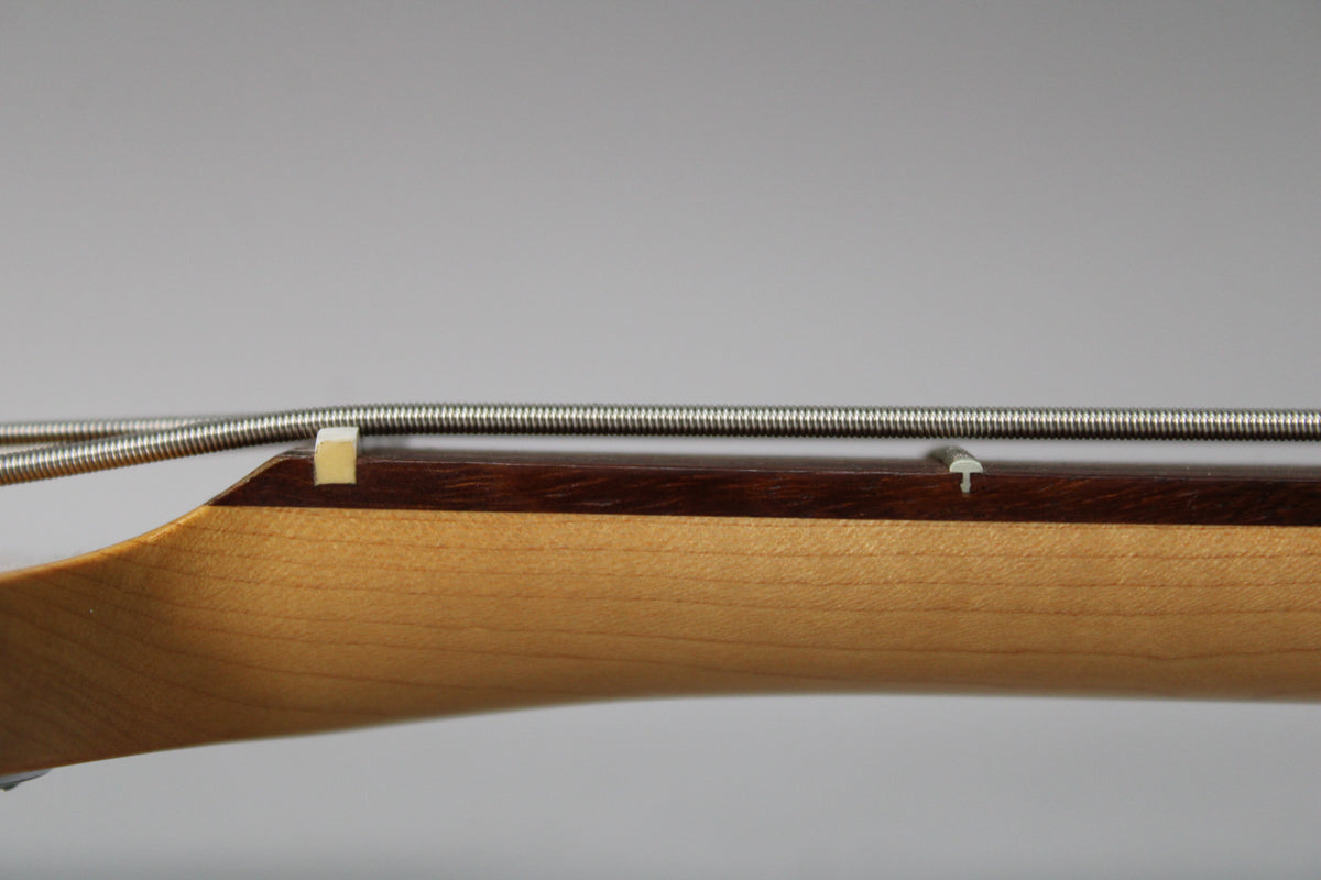 Close-up of the bass neck from the side, showing the light wood neck, dark fretboard, nut, and a single string tree.