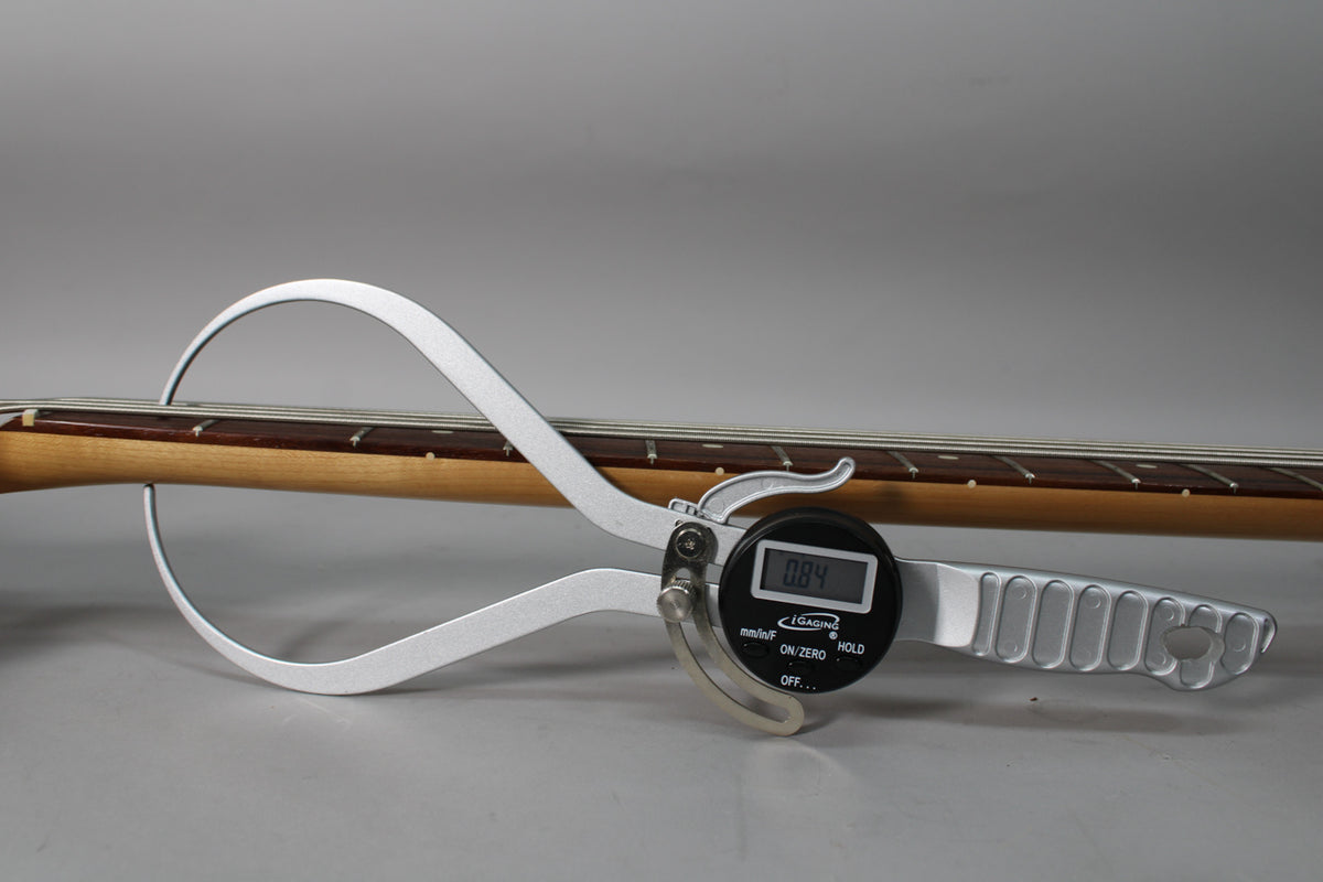 A digital caliper measures the bass's neck relief, showing "0.84" on its screen, with the fretboard and strings visible.