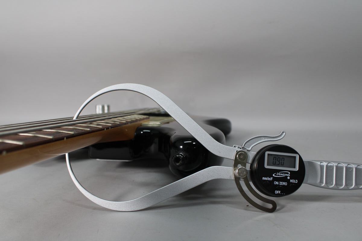 A digital caliper measures the neck relief of a black bass guitar, displaying "0.90".