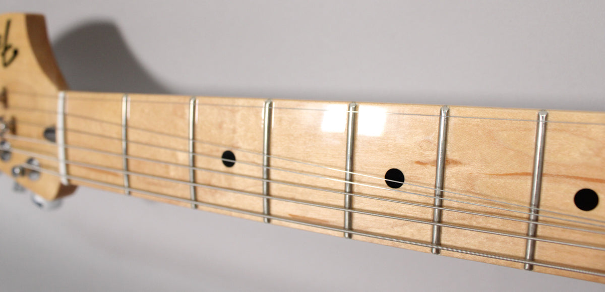 Close-up of the maple fretboard with a glossy finish, showing the first five frets, black dot inlays, and a partial view of the headstock.