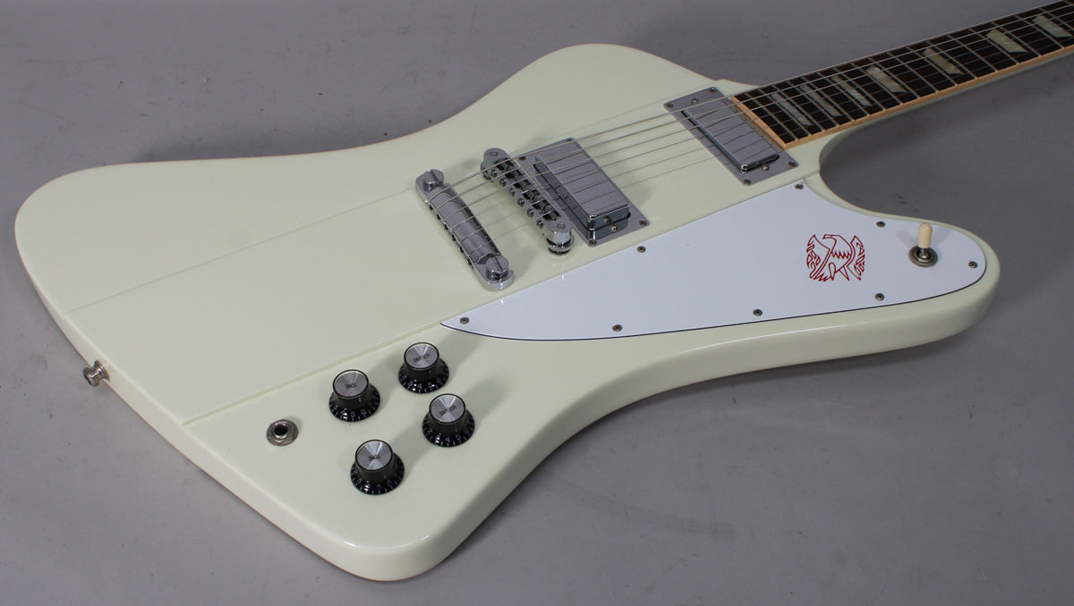 The classic white Firebird body, angled from the lower right, features a white pickguard with a red bird graphic, chrome hardware, and four black control knobs.