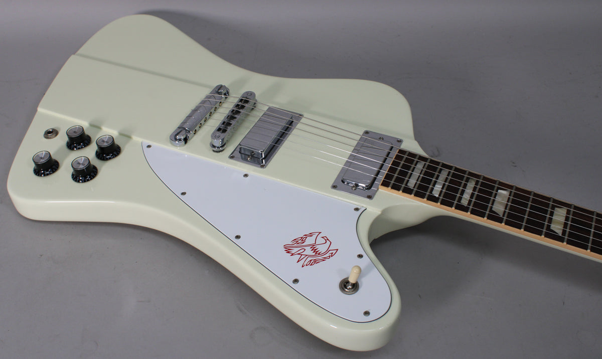 Classic white Firebird body, angled from above, shows four black control knobs, a white pickguard with red bird graphic, and two chrome humbuckers.