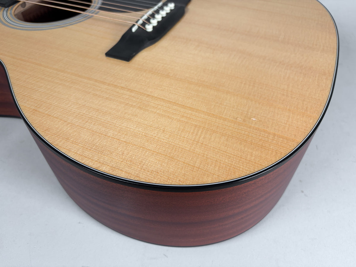 Upper bout of a natural finish acoustic guitar, showing the light wood grain top, dark sides, and a small ding near the lower edge.