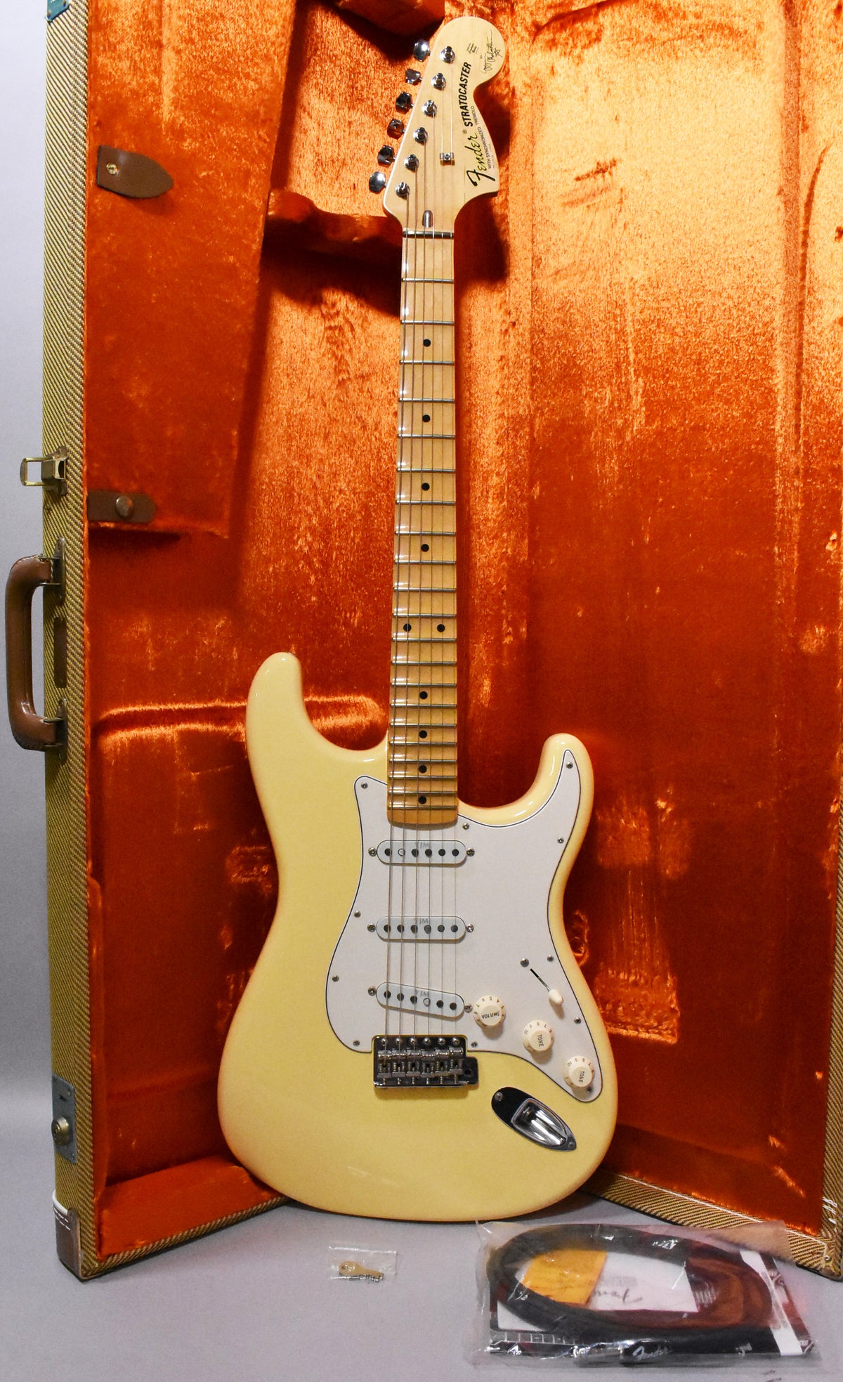 Full-length view of a vintage white Stratocaster with a maple neck, white pickguard, three single-coil pickups, and a tremolo bridge, standing in its tweed hard case.