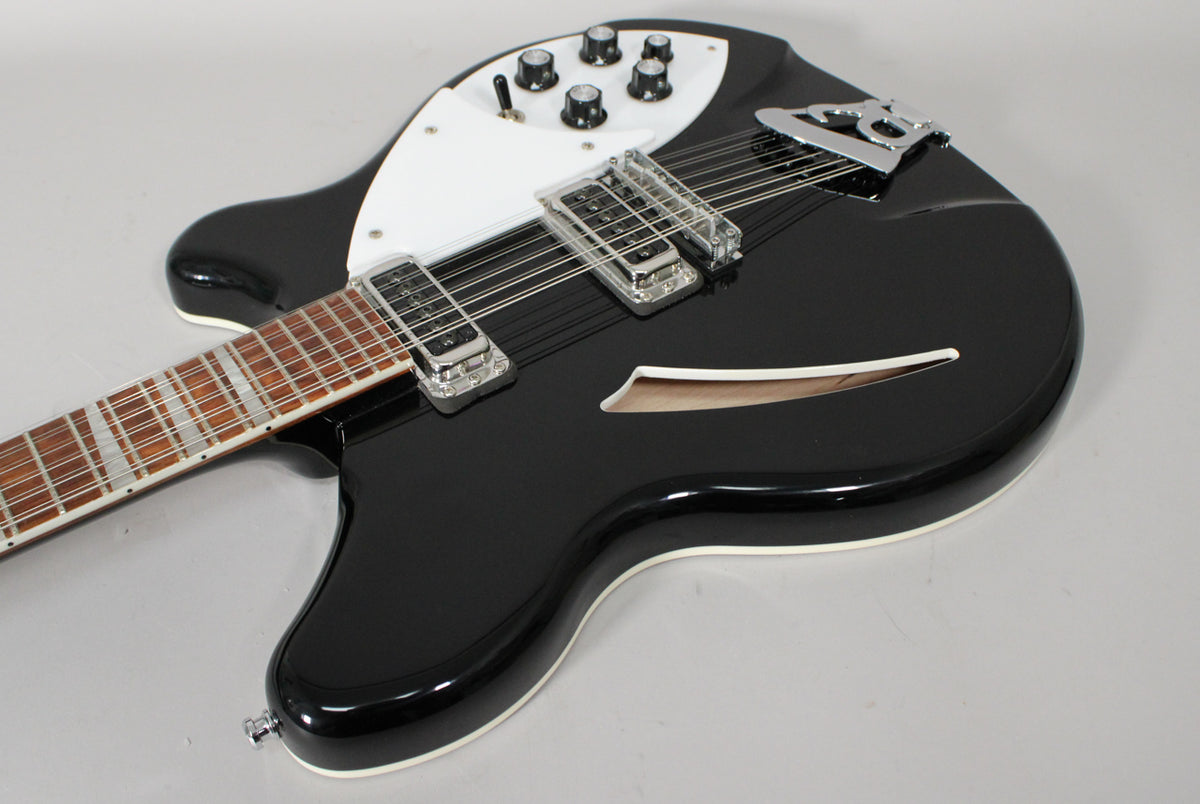 The glossy black 12-string electric guitar is angled, showing its white pickguard, five control knobs, two pickups, and f-hole.