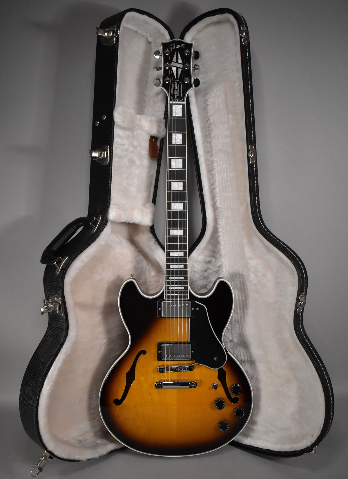 Full-length view of a glossy sunburst semi-hollow guitar with f-holes, black pickguard, chrome hardware, and a black headstock, standing in its open, plush-lined hard case.
