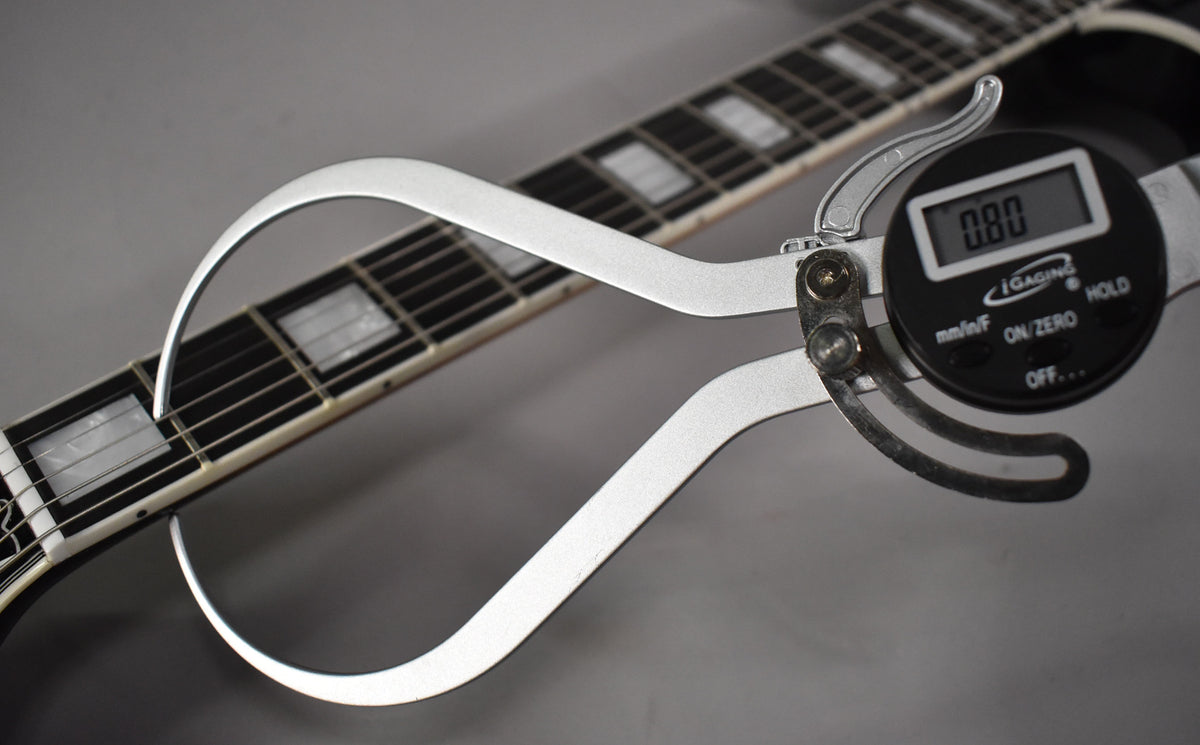 A digital caliper measures the guitar's fret height, showing 0.80mm, with the dark fretboard and block inlays visible.