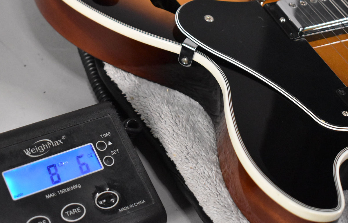 The guitar's lower bout in vintage sunburst is on a scale displaying 8 lbs 6 oz, with a black pickguard and cream binding.