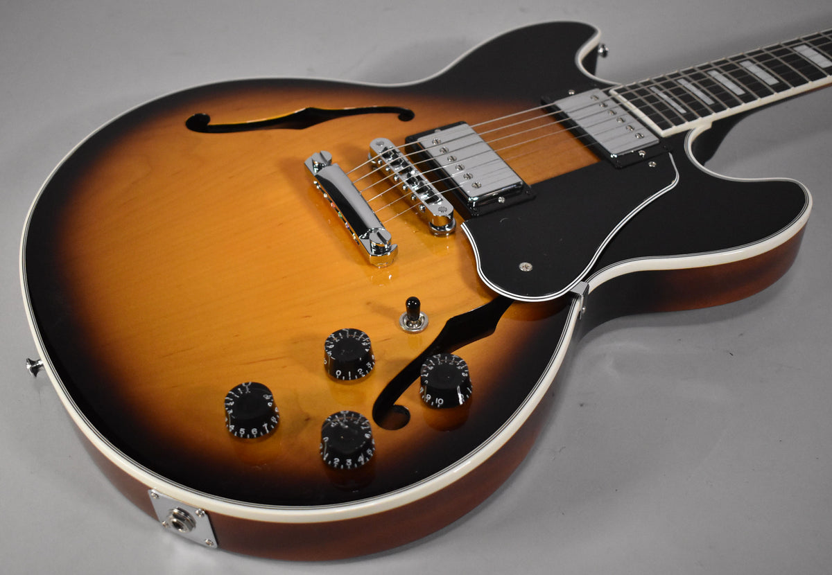 Close-up of a semi-hollow guitar in vintage sunburst, showing chrome humbuckers, black pickguard, f-holes, and four black control knobs.