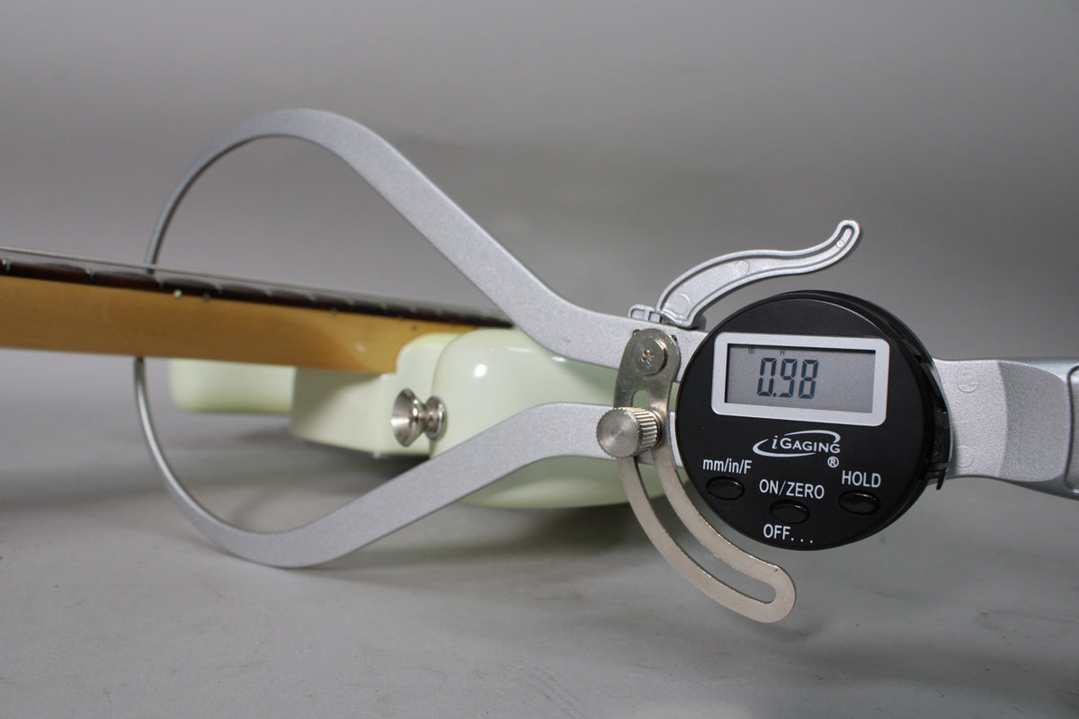 A digital caliper measures the Olympic White guitar's neck thickness at 0.98 inches, showing the fretboard and strap button.